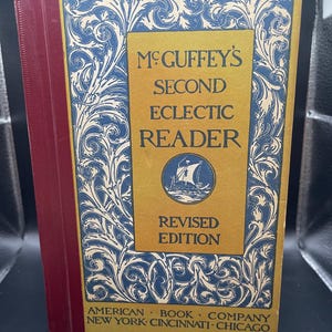 May include: A vintage copy of McGuffey's Second Eclectic Reader, Revised Edition. The book has a gold cover with blue and white decorative scrollwork. The spine is red. The book's title and publisher's information are visible.
