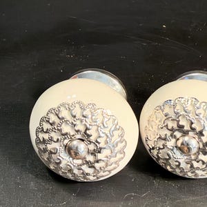 May include: Two round cabinet knobs with a smooth off-white ceramic surface. Each knob features a decorative silver-toned metal filigree overlay centered on the front, topped with a small polished silver-toned metal stud at the very center.