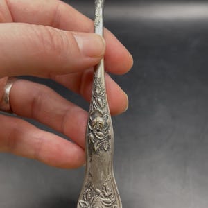 May include: A silver fork with three tines, featuring a floral rose design on the handle. The fork is held against a dark background, showcasing the intricate details of the embossed roses.
