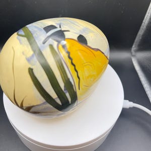 May include: A hand-painted, oval-shaped decorative egg. The egg features a yellow, black, and green design with a black hat. The egg is resting on a white, circular stand.