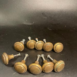 May include: A collection of ten round cabinet knobs with a weathered brass finish. Each knob features a central circular face with a raised border and a threaded metal screw extending from the back for installation on furniture or cabinetry doors.