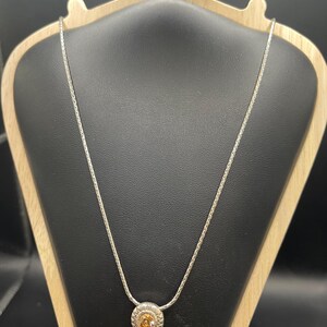 Op de afbeelding: Een zilveren ketting met een ronde hanger met een centrale oranje edelsteen, omgeven door een gedetailleerde zilveren zetting. De ketting wordt getoond op een zwarte sieradenstandaard met een houten frame, waarbij het ontwerp wordt benadrukt.