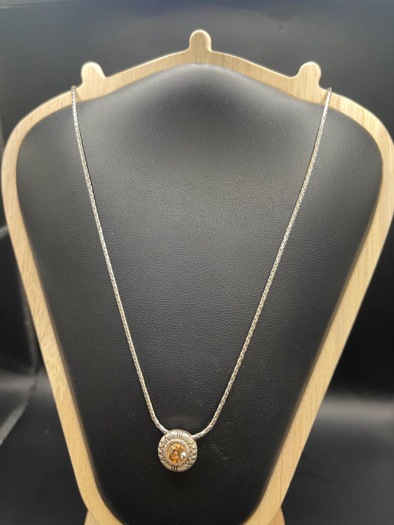 Op de afbeelding: Een zilveren ketting met een ronde hanger met een centrale oranje edelsteen, omgeven door een gedetailleerde zilveren zetting. De ketting wordt getoond op een zwarte sieradenstandaard met een houten frame, waarbij het ontwerp wordt benadrukt.