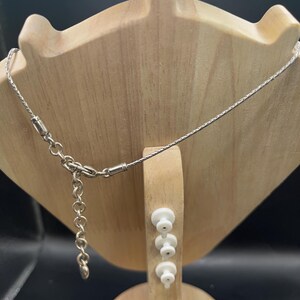 Op de afbeelding: Een zilverkleurige ketting met een fijne ketting wordt getoond op een houten sieradenstandaard. De ketting heeft een kleine sluiting en een verstelbare ketting. De standaard is lichtbruin en heeft een unieke vorm.