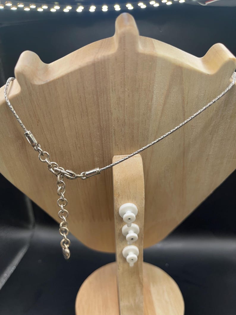 Op de afbeelding: Een zilverkleurige ketting met een fijne ketting wordt getoond op een houten sieradenstandaard. De ketting heeft een kleine sluiting en een verstelbare ketting. De standaard is lichtbruin en heeft een unieke vorm.