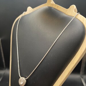 Op de afbeelding: Een zilveren ketting met een cirkelvormige hanger met een gedetailleerd ontwerp. De hanger is gecentreerd op een delicate ketting, gepresenteerd op een zwarte sieradenstandaard met een houten frame. De achtergrond is donker.