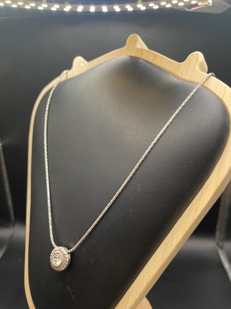 Op de afbeelding: Een zilveren ketting met een cirkelvormige hanger met een gedetailleerd ontwerp. De hanger is gecentreerd op een delicate ketting, gepresenteerd op een zwarte sieradenstandaard met een houten frame. De achtergrond is donker.