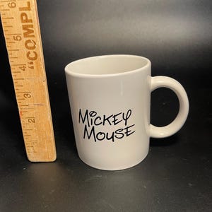 May include: A white ceramic mug with a curved handle. The mug features the text "Mickey Mouse" in black script. A wooden ruler is visible on the left side of the image.