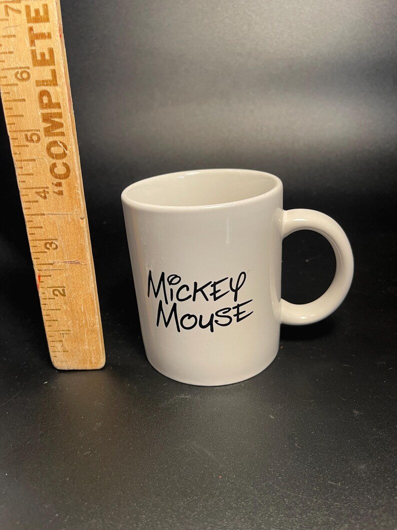 May include: A white ceramic mug with a curved handle. The mug features the text "Mickey Mouse" in black script. A wooden ruler is visible on the left side of the image.