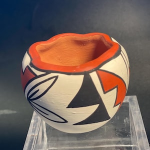 May include: A small, round, hand-painted clay pot featuring a white base with bold geometric patterns in black and terracotta orange. The rim is painted in a solid terracotta orange, while the interior reveals the natural, unglazed clay texture.