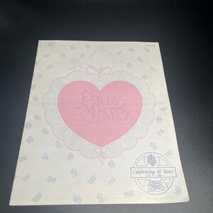 May include: A rectangular card with a pink heart-shaped design in the center, featuring the words "Precious Moments" in cursive. The card is decorated with small baby-themed illustrations and a logo that says "Celebrating 10 Years" and "Precious Moments".