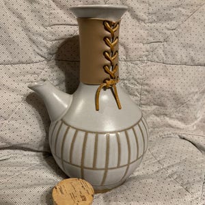 May include: A ceramic pitcher with a light gray glaze, featuring a tan leather wrap with a laced design. The pitcher has a spout and a rounded base with vertical lines. A cork stopper sits beside it.