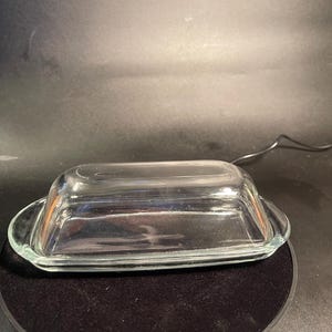 May include: A clear glass butter dish with a rectangular base and a matching domed lid. The dish features smooth, transparent glass construction with small handles on each end of the base, designed to hold a standard stick of butter securely.