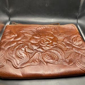 May include: A brown leather clutch with a floral rose design. The clutch features a zipper closure and a small handle. The embossed roses are the focal point of the design, adding a touch of elegance to the accessory.