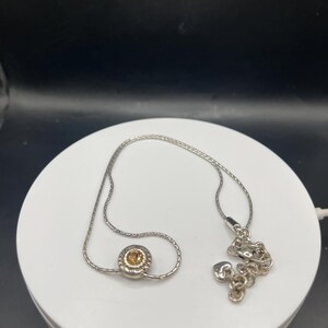 Op de afbeelding: Een zilveren ketting met een kleine, ronde hanger met een centrale amberkleurige steen. De ketting wordt gepresenteerd op een wit oppervlak tegen een donkere achtergrond. De ketting is delicaat en de hanger is het middelpunt.