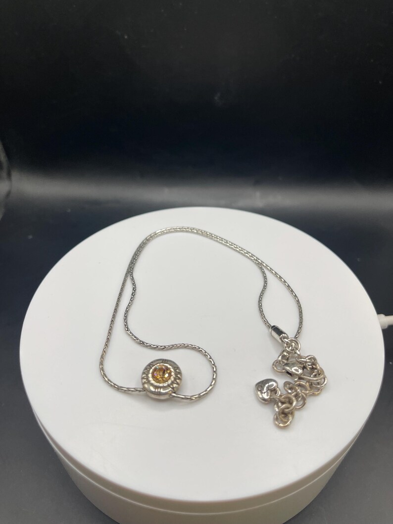 Op de afbeelding: Een zilveren ketting met een kleine, ronde hanger met een centrale amberkleurige steen. De ketting wordt gepresenteerd op een wit oppervlak tegen een donkere achtergrond. De ketting is delicaat en de hanger is het middelpunt.