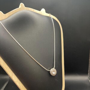 Op de afbeelding: Een zilveren ketting met een kleine, ronde hanger. De hanger heeft een textuurontwerp rond een centrale heldere steen. De ketting wordt gepresenteerd op een zwarte sieradenstandaard.