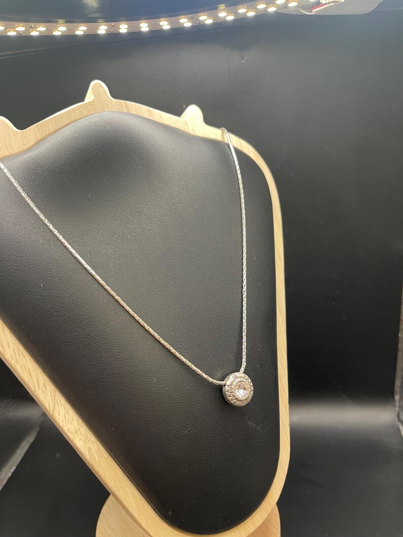 Op de afbeelding: Een zilveren ketting met een kleine, ronde hanger. De hanger heeft een textuurontwerp rond een centrale heldere steen. De ketting wordt gepresenteerd op een zwarte sieradenstandaard.