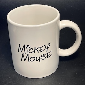 May include: A white ceramic mug with a curved handle. The words "Mickey Mouse" are printed in black script on the side of the mug. The mug is empty and sits on a dark surface.