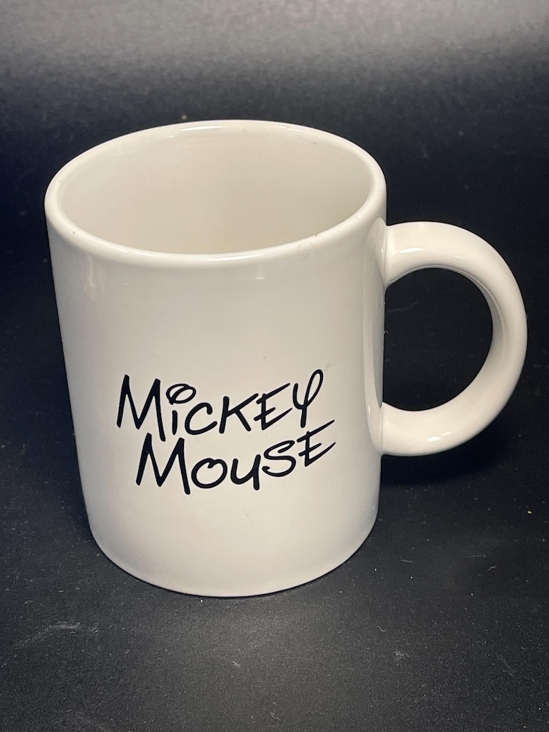 May include: A white ceramic mug with a curved handle. The words "Mickey Mouse" are printed in black script on the side of the mug. The mug is empty and sits on a dark surface.