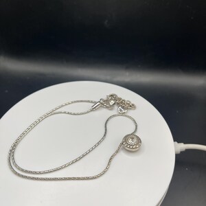 Op de afbeelding: Een zilveren ketting met een fijne ketting en een ronde hanger. De hanger heeft een centrale heldere steen omringd door kleinere stenen, wat een sprankelend effect cre&euml;ert. De ketting wordt gepresenteerd op een wit oppervlak tegen een zwarte achtergrond.