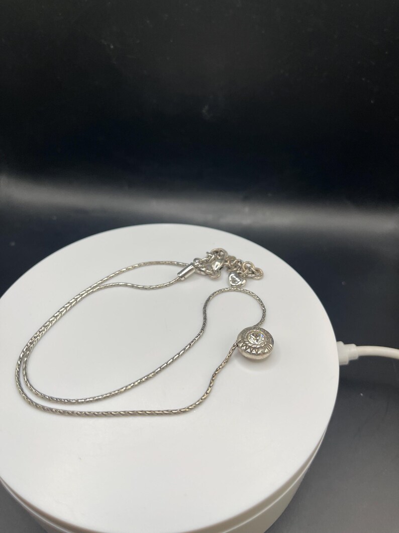 Op de afbeelding: Een zilveren ketting met een fijne ketting en een ronde hanger. De hanger heeft een centrale heldere steen omringd door kleinere stenen, wat een sprankelend effect cre&euml;ert. De ketting wordt gepresenteerd op een wit oppervlak tegen een zwarte achtergrond.