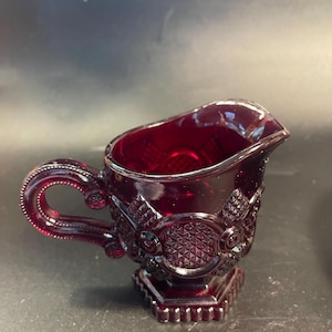 May include: A deep red glass creamer pitcher featuring a textured diamond pattern and circular embossed details. The vessel has a curved handle with a beaded edge, a small pouring spout, and a hexagonal base with a scalloped bottom rim.