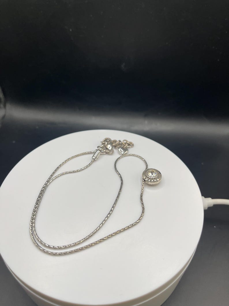 Op de afbeelding: Een zilverkleurige ketting met een fijne ketting. De ketting heeft een ronde hanger met een centrale heldere steen. De ketting is ongeveer 40 cm lang.