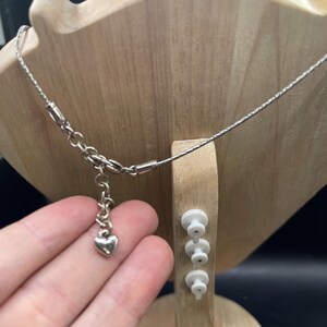 Op de afbeelding: Een zilveren ketting met een hartvormige hanger wordt getoond op een houten sieradenstandaard. De ketting is delicaat, met een fijne ketting en een kleine hartvormige bedel. De standaard is lichtbruin en heeft een unieke vorm.