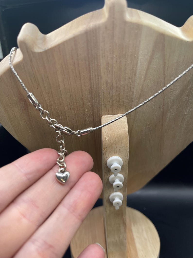 Op de afbeelding: Een zilveren ketting met een hartvormige hanger wordt getoond op een houten sieradenstandaard. De ketting is delicaat, met een fijne ketting en een kleine hartvormige bedel. De standaard is lichtbruin en heeft een unieke vorm.