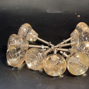 May include: A collection of round cabinet knobs made of clear resin with embedded silver-toned metallic fibers. Each knob features a smooth, spherical head attached to a threaded metal bolt with a small hexagonal nut for secure installation.