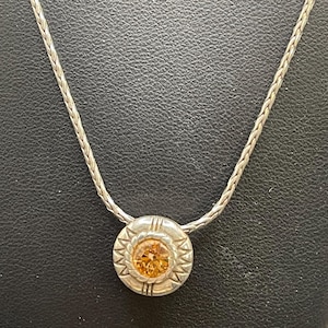 Op de afbeelding: Zilverkleurige ketting met een cirkelvormige hanger met een oranje edelsteen in het midden. De hanger heeft een decoratieve rand met een zonnesterontwerp. De ketting heeft een geweven stijl.