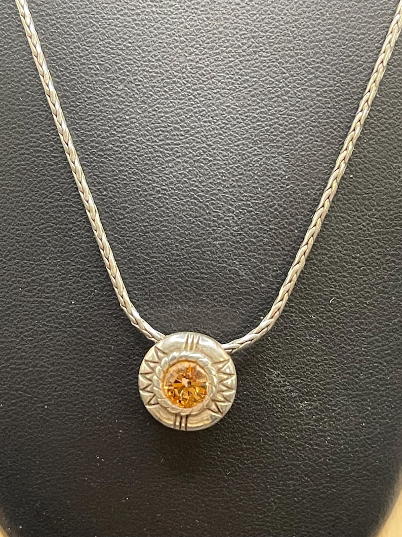 Op de afbeelding: Zilverkleurige ketting met een cirkelvormige hanger met een oranje edelsteen in het midden. De hanger heeft een decoratieve rand met een zonnesterontwerp. De ketting heeft een geweven stijl.