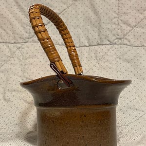 May include: A brown ceramic basket with a woven handle. The basket has a cylindrical body with a slightly flared rim. The handle is made of woven, light brown material and is attached to the basket with metal hardware. The basket has a rustic, handmade appearance.