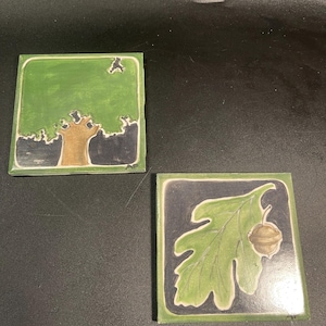 May include: Two square ceramic tiles featuring nature-inspired illustrations. One tile depicts a stylized tree with a green canopy and brown trunk. The other tile shows a green oak leaf and a small brown acorn against a dark blue background.