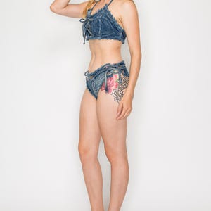 May include: A denim two-piece set comprising a halter top and low-rise shorts. The top has tie closures at the front, and the shorts have tie details at the sides. The set is made of blue denim fabric. The model is wearing clear heeled sandals.