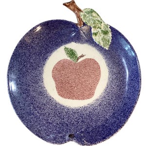 May include: A ceramic apple-shaped serving plate. The plate has a speckled blue exterior with a white interior circle. A pink apple design is in the centre. A brown stem and green leaf are attached to the top.