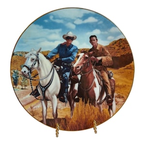 Puede incluir: Un plato decorativo redondo de colección con una ilustración colorida de dos jinetes a caballo en un paisaje desértico. Un jinete lleva un sombrero blanco y camisa azul sobre un caballo blanco, el otro una chaqueta de flecos beige sobre un caballo.
