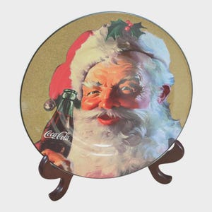 Puede incluir: Un plato decorativo con Santa Claus sosteniendo una botella de Coca-Cola. El plato tiene un fondo dorado y un soporte marrón. Santa lleva un gorro rojo y blanco y tiene una larga barba blanca. La botella de Coca-Cola es verde y roja.