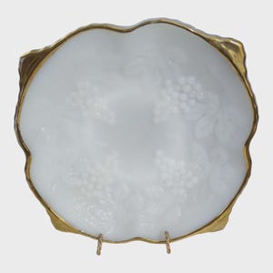 May include: A white, decorative serving dish with a gold-colored rim. The dish features a raised grape and leaf design. The dish is supported by two gold-colored metal stands. The dish is likely made of glass or ceramic.