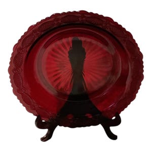 Puede incluir: Un plato de vidrio rojo rubí con un borde festoneado decorativo y un diseño central. El plato se exhibe sobre un soporte marrón oscuro, mostrando sus intrincados detalles y su color vibrante. Este plato de estilo vintage es perfecto para servir o exhibir.