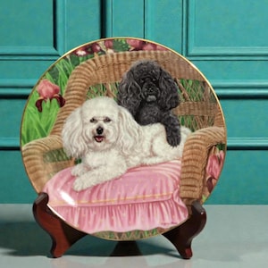 May include: Decorative plate featuring two dogs, a white Bichon Frise and a black Poodle, resting on a pink cushion within a wicker chair. The plate has a floral border and is displayed on a wooden stand.