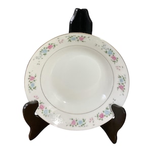 May include: A white ceramic plate with a gold rim and a floral pattern of pink and blue roses. The plate is displayed on a dark brown wooden stand, showcasing its elegant design. This decorative plate is perfect for serving meals or as a display piece.