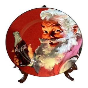 Puede incluir: Plato decorativo con Papá Noel sosteniendo una botella de Coca-Cola. El plato tiene un fondo rojo con una ilustración vintage de Papá Noel con traje rojo y barba blanca. El plato se exhibe en un soporte marrón.