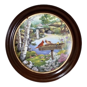 Puede incluir: Un plato decorativo redondo con un marco de madera marrón. El plato presenta una pintura detallada de una escena al aire libre serena con un estanque, árboles y varios pájaros. La obra de arte incluye un par de cardenales rojos y otras aves en un comedero.