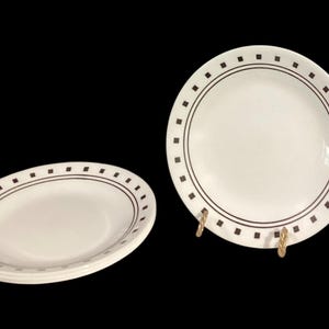 May include: White ceramic plates with a brown square and line design around the rim. One plate is oval, and two round plates are visible, one on a gold stand. The plates are on a black background.