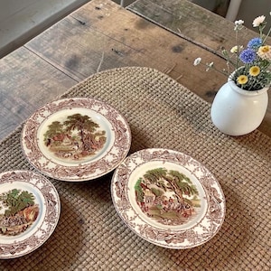 May include: Three antique decorative plates with a brown and white pastoral scene. The plates are arranged on a woven mat on a wooden table. A small white vase with colorful flowers sits nearby.