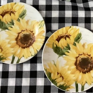 May include: Two oval-shaped plates with a sunflower design. The plates feature a white background with yellow sunflowers, green leaves, and brown centers. The plates are set on a black and white checkered tablecloth.
