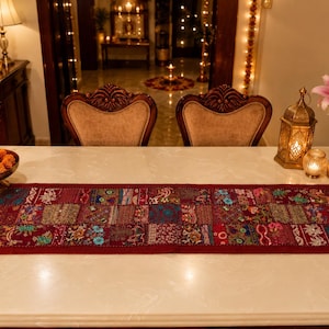 May include: A rectangular, patchwork-style table runner in shades of burgundy, red, and various other colors. The runner is decorated with floral and geometric patterns. The runner is placed on a white table, with candles and flowers in the background.