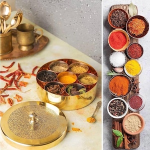 May include: A gold-colored spice container with a decorative lid, filled with various spices. The container is round with multiple compartments. A wooden board displays small bowls of spices, including saffron, red pepper, and cinnamon sticks. A spoon rests near the container.